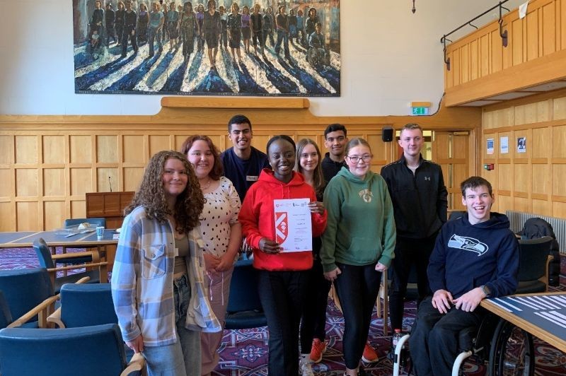nine smiling young people in casual clothes, one of them sitting in a wheelchair, in a wood panelled room with blue patterned carpet